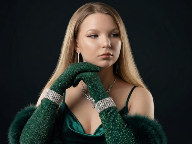 MariaRobinson with blonde hair, green glitter gloves, and a green satin dress, wearing a silver necklace and earrings, touching her chin with a serious expression.