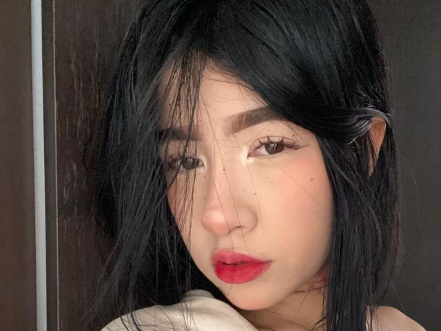 SophiaXSweety with black hair, red lips, and light skin, looking at the camera with half-closed eyes, her nose slightly shiny, and a small mole on her cheek.