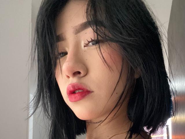 SophiaXSweety has black, shoulder-length hair, red lips, and is looking down with slightly parted pink lips. She has dark eyebrows, eyelashes, and a small nose.