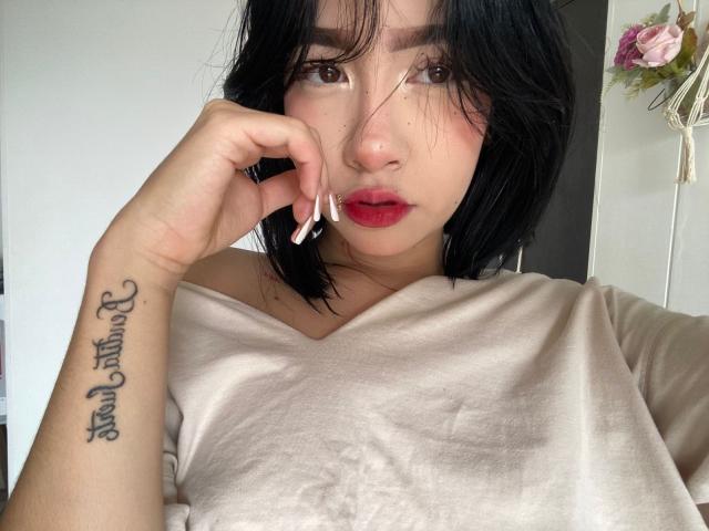 SophiaXSweety with black hair, red lipstick, and a tattoo on her left forearm, wearing a beige shirt, touching her nose with white-painted nails. Her tits are partially visible.