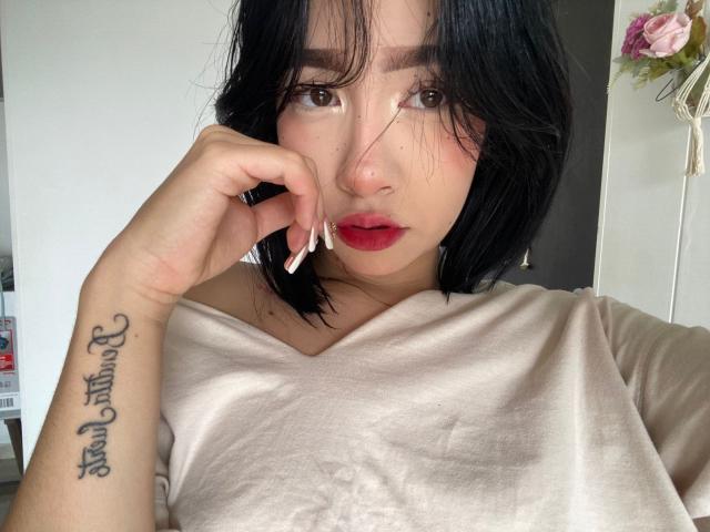 SophiaXSweety with black hair, red lipstick, and a tattoo on her right forearm, wearing a beige shirt, touching her cheek with long, white-tipped nails.