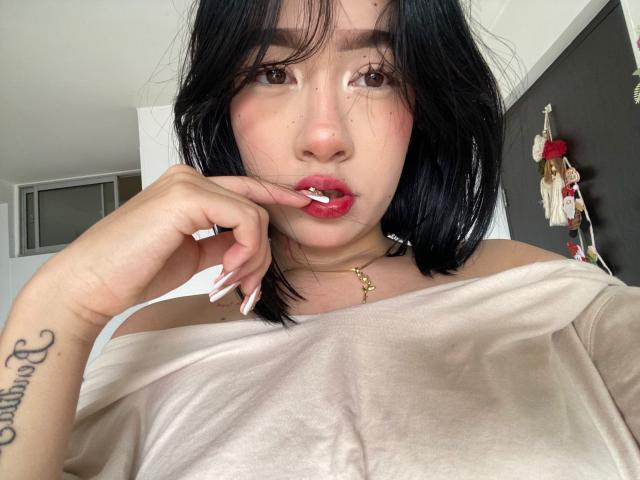 SophiaXSweety with black hair, pink lipstick, and a gold necklace, touching her lip with a finger, wearing a beige off-shoulder top, showing a tattoo on her wrist.