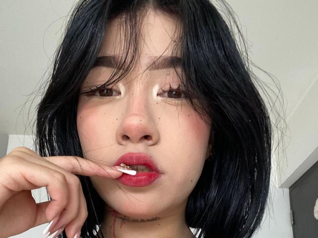 SophiaXSweety has black hair, red lipstick, and is biting her white-manicured nail with a slightly open mouth, showing her pink tongue and a small cut below her chin.