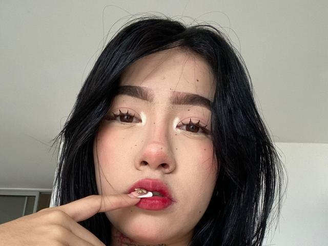 SophiaXSweety with black hair, brown eyes, and red lips, biting her finger with long, fake eyelashes, and visible moles on her face, in a selfie.