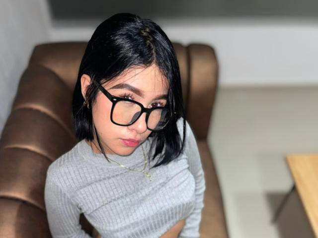 SophiaXSweety with black glasses, grey ribbed top, and black bob, looking down, sitting on brown leather couch, small tits, cute, light brown skin, gold necklace.