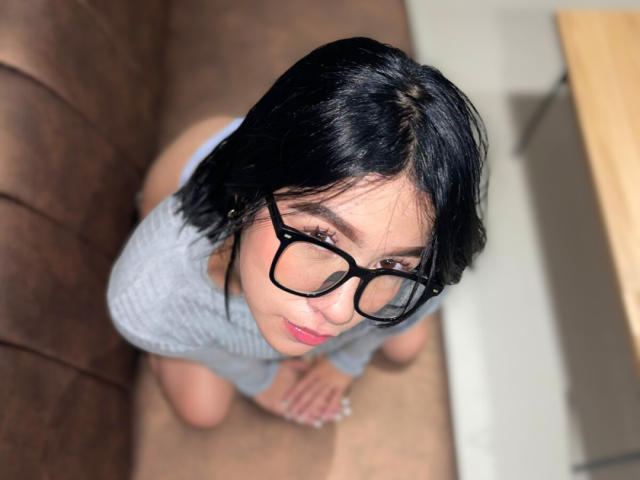 SophiaXSweety with black glasses, short black hair, and pink lipstick, kneeling on a brown floor, wearing a gray top, looking up with a slight smile.