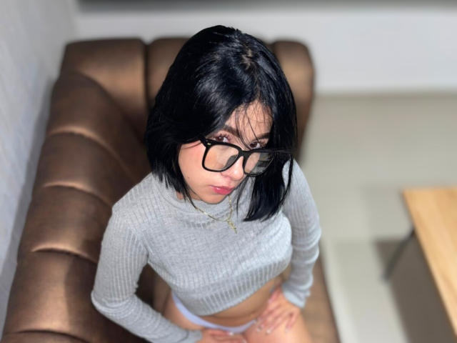 SophiaXSweety with black bob, glasses, grey ribbed crop top, midriff, hands on brown leather bench, looking up, small tits, black hair, light skin, subtle makeup, necklace.