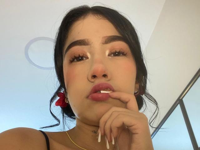 SophiaXSweety with dark hair, red scrunchie, nose ring, and long nails, biting her finger, looking up, has brown eyebrows, and wears a gold necklace.