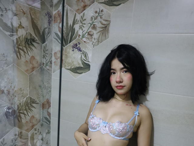 SophiaXSweety with short black hair, wearing a light blue lace bra with pink floral details, standing in a bathroom, showing her small tits and a tattoo on her right chest.