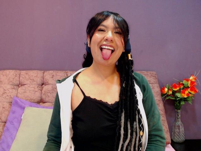 BeckyHuston with black braided hair, green cardigan, white vest, black tank top, sticking out tongue, smiling, medium tits, pinkish-brown couch, purple wall, vase with red flowers.
