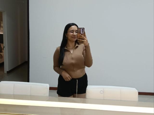 Karollteyes, com cabelo preto longo, óculos, blusa bege e shorts pretos, segurando um celular, selfie no espelho, mostrando seus pequenos seios, em um quarto moderno com cadeiras brancas..
