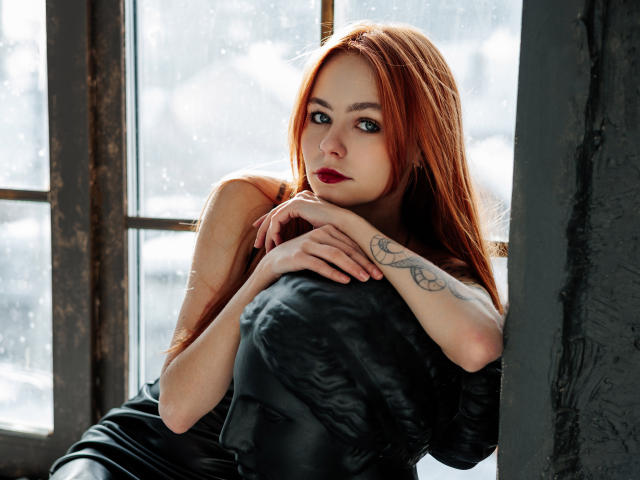 MiaCuteAngel with red hair, black dress, tattoo on right forearm, leaning on black pillow, hands on pillow, looking at camera, window behind her, soft light.