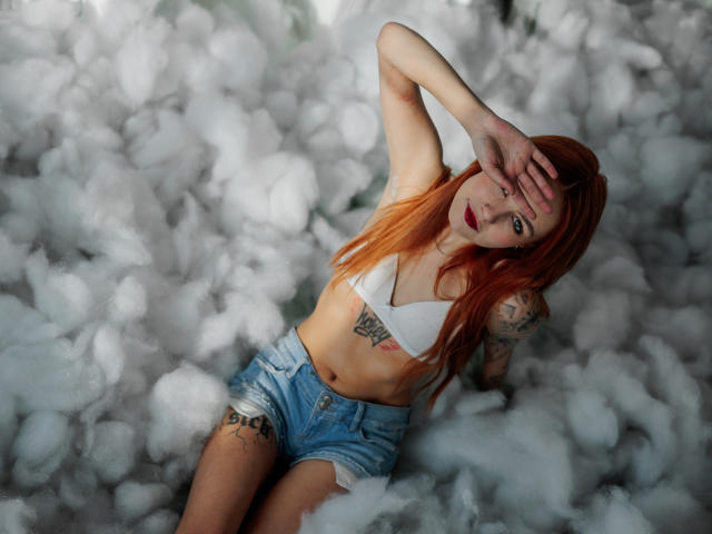 MiaCuteAngel, a red-haired woman with tattoos, sits in a cloud of white, fluffy material. She wears a white, triangular crop top and blue, distressed denim shorts. Her left arm covers her eyes, revealing a tattoo on her right forearm. Her right arm rests on her thigh, and she has another tattoo on her left thigh. Her long hair is orange at the tips. The image has a soft, almost dreamy quality with a focus on her relaxed, yet slightly defiant, posture. The texture of the fluffy material contrasts with her smooth, fair skin.