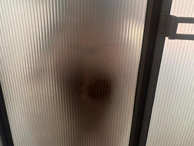 Close-up of a frosted glass door. The glass is slightly ribbed, creating vertical lines. A dark, circular stain is centered on the glass, slightly blurred due to the texture. The right edge shows a metal hinge. The glass is translucent, with light diffusing through the ribbed surface. The image focuses on the texture and stain, emphasizing the contrast between the dark stain and the frosted glass.