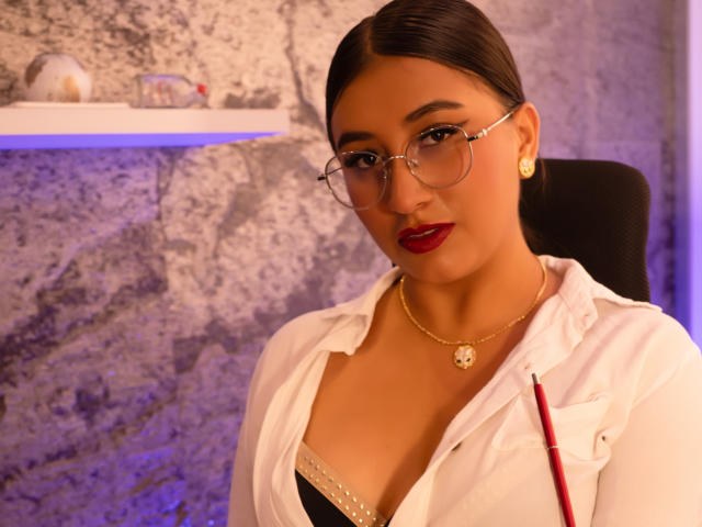 Catalina Rivero, con gli occhiali, il rossetto rosso e una collana d'oro, indossa una camicia bianca con una penna attaccata al petto. Ha un seno piccolo e un viso carino..