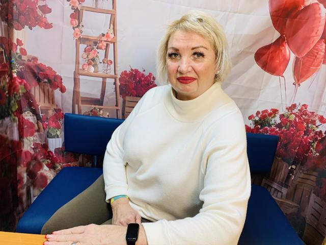 RenataDaster with short blonde hair, red lipstick, white sweater, and black watch, sitting on blue chair, smiling, wearing tan pants, red balloons in background.