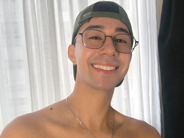 OliverHot69, torse nu, portant une casquette verte et des lunettes, souriant, avec un visage rasé de près, un collier en chaîne et une petite tache de naissance sur le côté gauche de son cou..