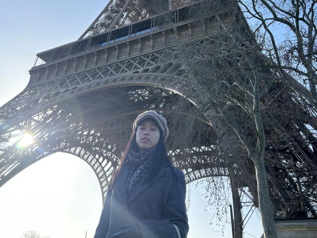 NashiroSakuraii stands in front of the Eiffel Tower, wearing a gray hat, black scarf, and coat, with a sun flare and leafless trees in the background.