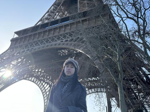 NashiroSakuraii, with a hat and dark coat, stands in front of the Eiffel Tower, her face partially in shadow, sunlight behind her, bare trees to the right.