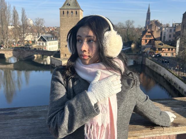 NashiroSakuraii with long black hair, wearing fluffy white ear warmers, gray coat, pink scarf, and white gloves, stands by a wooden railing, looking pensive. Sunlit, wintery cityscape.