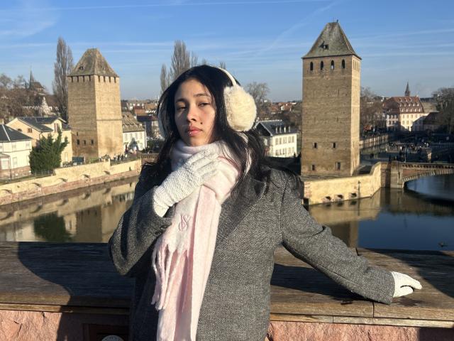 NashiroSakuraii stands on a wooden railing, wearing a gray coat, pink scarf, white gloves, and fluffy ear warmers, with her hand near her mouth, looking serious.