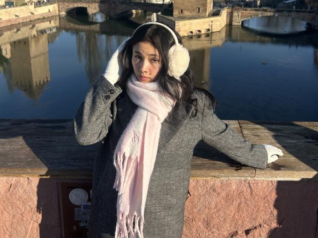 NashiroSakuraii with dark hair, wearing a gray coat, pink scarf, white earmuffs, and white gloves, leaning on a wooden railing, hand in hair, standing by a reflective waterway.