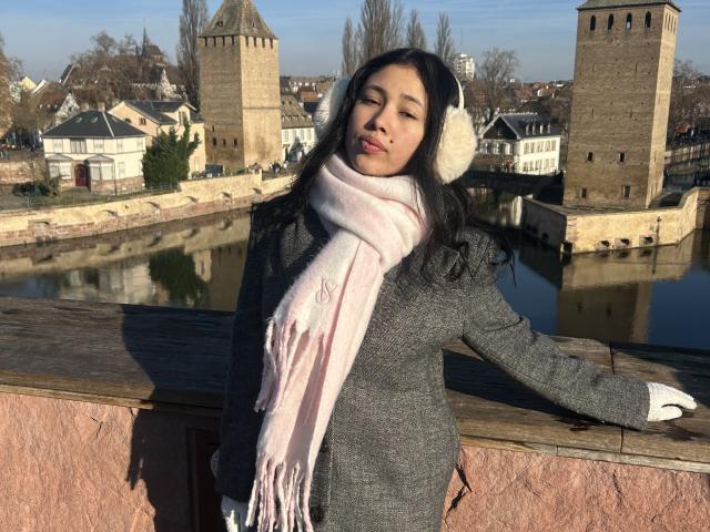 NashiroSakuraii stands in front of a river, wearing a gray coat, pink scarf, white earmuffs, and white gloves, with a slightly smug expression, eyes half-closed.
