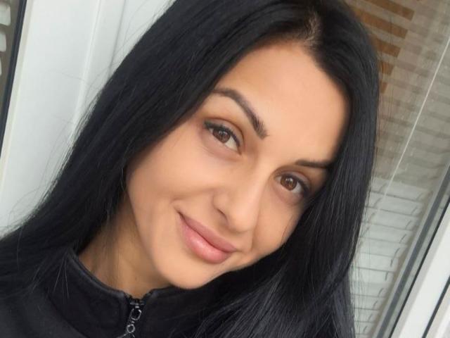 EvaClarkX has straight, black hair falling over her shoulders, brown eyes with subtle eyeliner, and a light skin tone. She wears a black zip-up top. Her lips are slightly parted in a gentle smile. The image is a close-up, focusing on her face, with a white window frame on the left and a brick wall on the right. The lighting is even, highlighting her natural features.