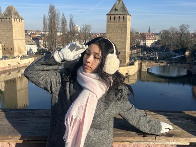 NashiroSakuraii with puffy white earphones, gray sweater, pink scarf, white gloves, and long dark hair, standing by a wooden railing, eyes closed, hand on ear.