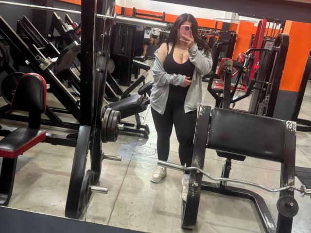 VenusxMusa mit großen Brüsten, in einem schwarzen Sport-BH, schwarzen Leggings und einer grauen Kapuzenjacke, macht ein Selfie vor einem Spiegel im Fitnessstudio und trägt weiße Turnschuhe..