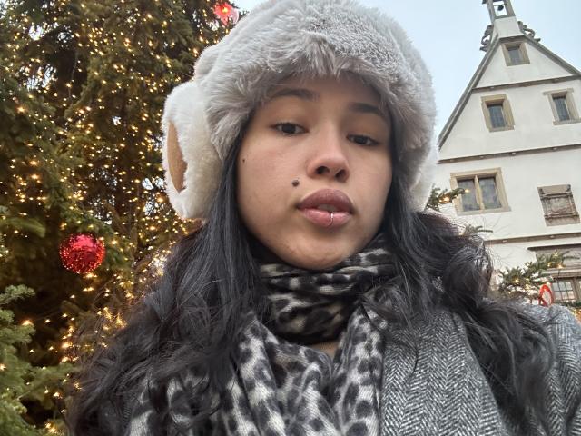 NashiroSakuraii with long black hair, pouty lips, gray fur hat, leopard scarf, and a mole on her right cheek, standing in front of a decorated Christmas tree.