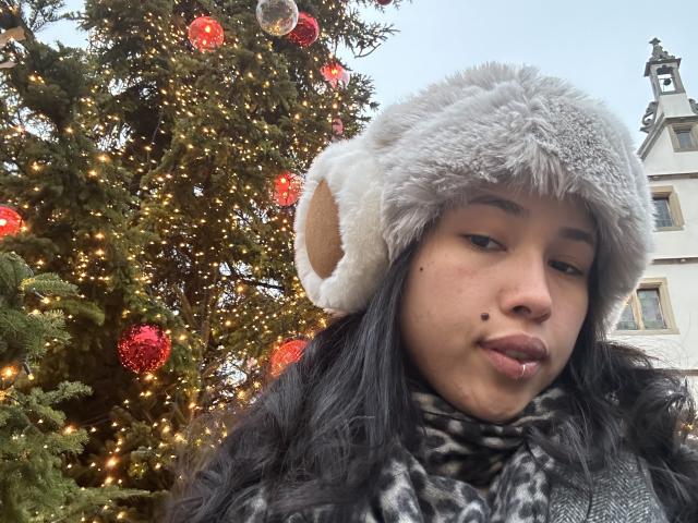NashiroSakuraii has a cute face, dark wavy hair, and is wearing a fluffy gray hat with a brown ear, looking down with a slight pout, in front of a Christmas tree.