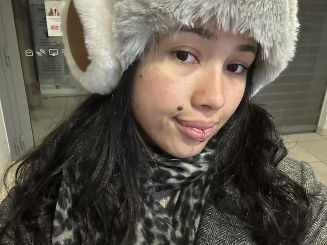 NashiroSakuraii with a grey fur hat, black hair, and a cute lip piercing, sticking out her tongue, wearing a leopard print scarf, in a selfie.