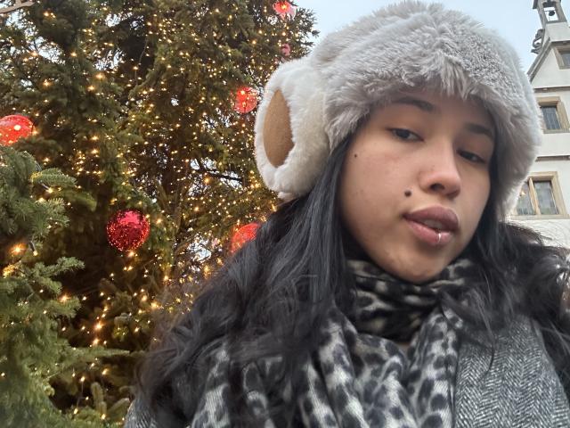 NashiroSakuraii has dark hair, a gray furry hat with a bear ear, a leopard print scarf, and a small mole on her right cheek. Christmas tree in background.