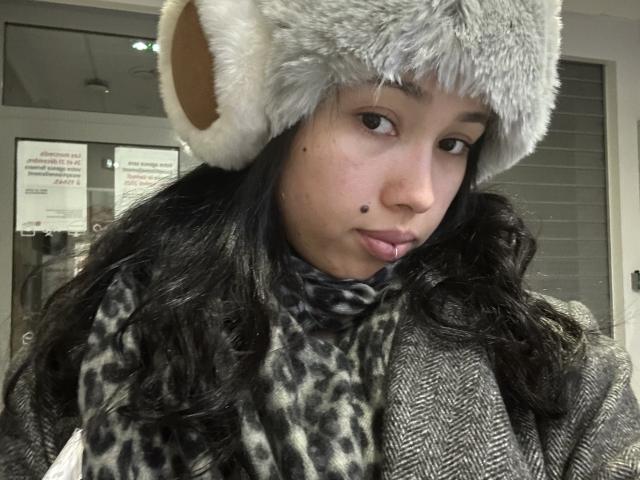 NashiroSakuraii with a gray furry bear hat, leopard scarf, and houndstooth coat, has a small beauty mark on her left cheek, and long wavy black hair.