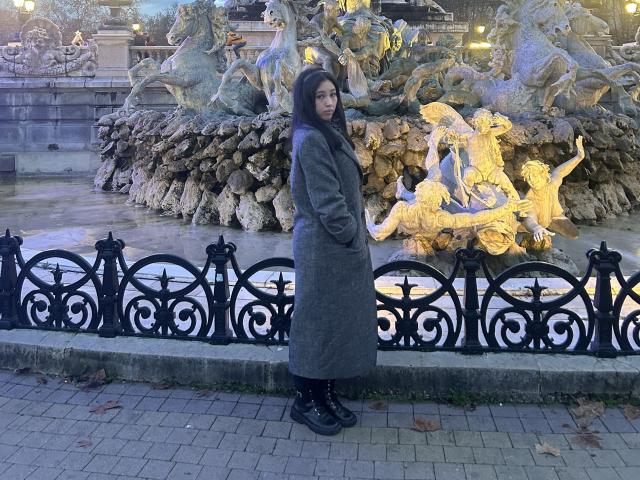 NashiroSakuraii with long black hair, wearing a grey coat and black boots, stands in front of a golden, detailed fountain, looking over her shoulder, in a cobblestone area.