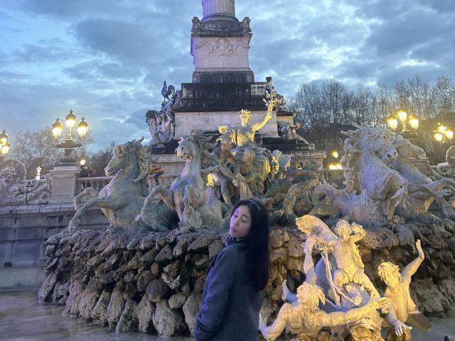 NashiroSakuraii with long dark hair stands in front of a massive, detailed stone fountain featuring mythical, erect horses and nymphs. She's wearing a dark jacket, eyes closed.