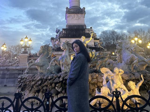 NashiroSakuraii stands in front of a statue with horses and mythical creatures, wearing a long gray coat, looking over her shoulder with dark hair and a serious expression.