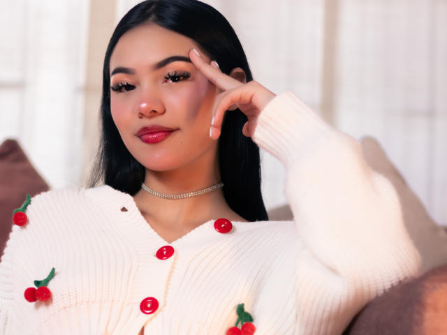 ValerieCasanova with long black hair, red lipstick, and white knitted cardigan with red cherries, wearing a choker, touching her cheek, and looking at the camera.