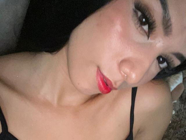 AnieCartier's face, showing dark eyebrows, long eyelashes, red lipstick, and a slight nose piercing, lying with black straps on her shoulders, in a close-up shot.