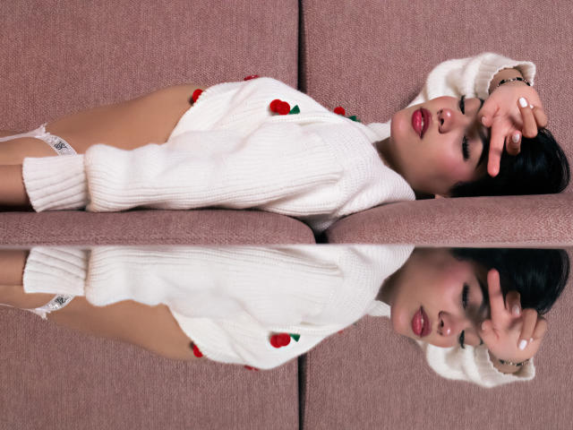 ValerieCasanova lying on a pink couch, wearing a white sweater with strawberry embroidery, white lace panties, and red lipstick, with her hand covering her face, reflected in a mirror.