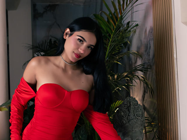 ValerieCasanova with long black hair, red strapless dress, and matching gloves, showing her tits, choker, and red lipstick, standing near a green plant and a carved stone.