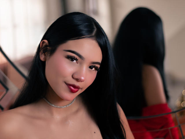 ValerieCasanova has long black hair, red lipstick, and a shiny skin, wearing a silver choker, looking at the camera with a slight smile, reflection in the background.