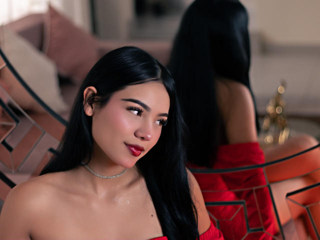 ValerieCasanova with long black hair, red dress, off-shoulder, shiny skin, subtle smile, looking to the side, reflection in mirror, gold necklace, blurred background, soft lighting.