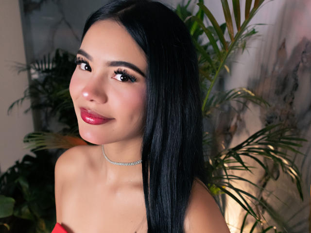 ValerieCasanova with long black hair, red lipstick, and a sparkling choker, smiling at the camera, showing her shiny, plump, glossy, red-ass lips.