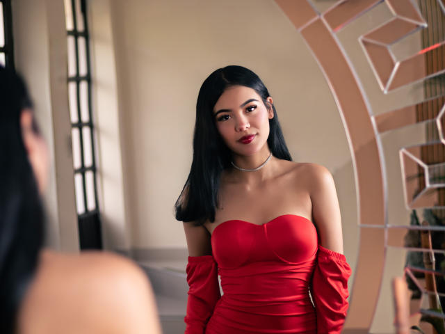 ValerieCasanova with long black hair, wearing a red strapless dress, looking in a mirror, showing her tits, choker necklace, and a subtle smile, in a stylish room.