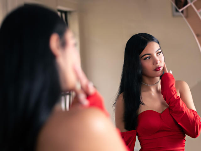 ValerieCasanova with black hair, red dress, and red gloves, looking at herself in a mirror, touching her chin, showing off her tits and smooth skin. She's wearing a necklace.