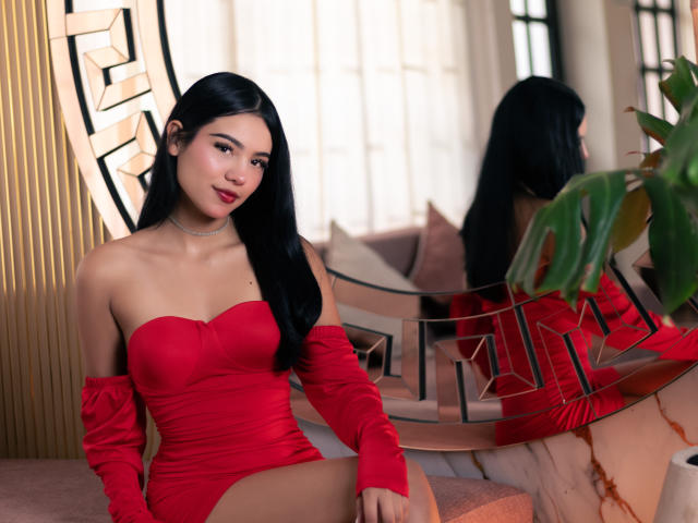 ValerieCasanova in a red, off-shoulder, ruched dress with matching gloves, sitting, showing her tits, black hair, red lips, and a reflection in a mirror.