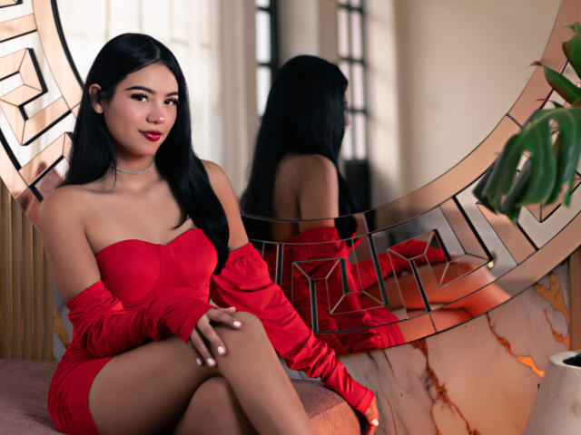 ValerieCasanova with long black hair, red off-shoulder dress, and matching gloves, sitting with legs crossed, showing her ass, in front of a mirrored wall, smiling.