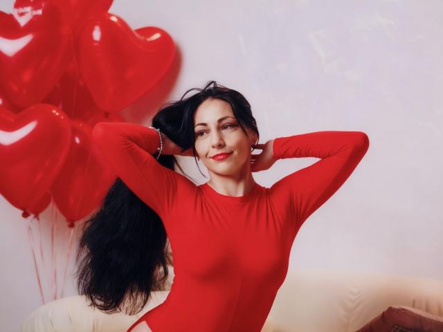 LiluGrants with black hair, wearing a red long-sleeve bodysuit, hands in hair, red heart balloons, sitting on a white couch, smiling, red lipstick, confident, sexy.
